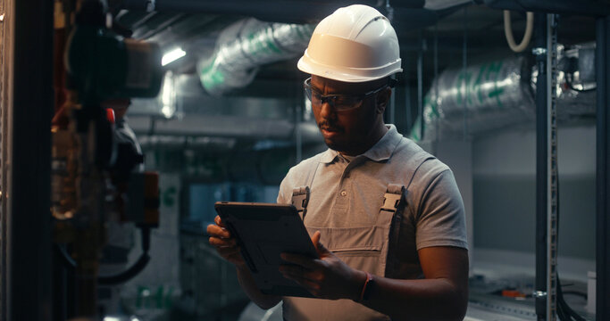 Male technician worker wearing safety uniform and hard hat works using tablet computer. African American inspector checks pipeline system on modern factory, plant or industrial energy facility.