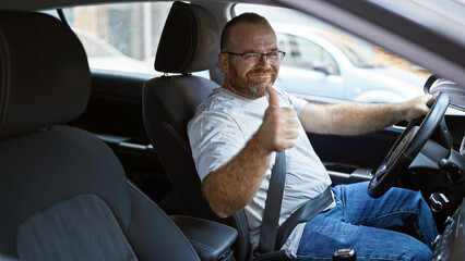 Cheery middle-aged caucasian man driving car down urban street, giving thumb up gesture and...