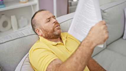 Handsome middle-aged caucasian man suffering in the hot weather at home, using a document as a...