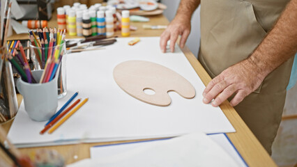 Handsome, middle-aged caucasian man artist, beard and glasses, passionately engaged in his hobby, painting a portrait on canvas, at an indoor art studio, holding palette and paintbrushes.