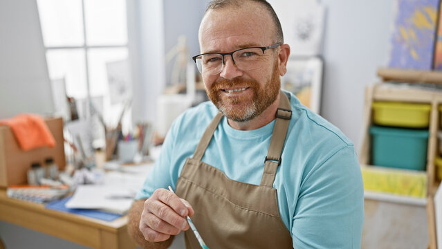 Attractive middle-aged caucasian man confidently enjoying his hobby, smiling as he draws a piece of art in his cozy studio, surrounded by paintbrushes and the aroma of creativity
