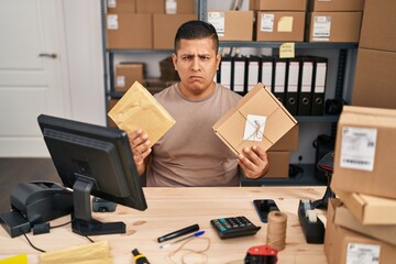 Hispanic young man working at small business ecommerce holding packages skeptic and nervous, frowning upset because of problem. negative person.