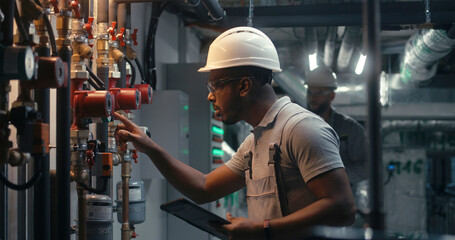 Male technician worker wearing safety uniform and hard hat works using tablet computer. African American inspector checks pipeline system on modern factory, plant or industrial energy facility. - obrazy, fototapety, plakaty
