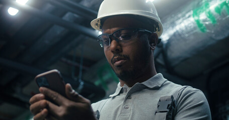 Close up of African American engineer using phone during break, surfing the Internet. Heavy industry worker in safety uniform and hard hat works on modern manufacturing factory or industrial plant.
