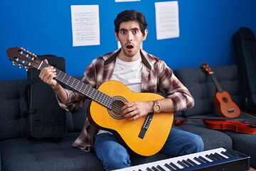 Young hispanic man playing classic guitar at music studio scared and amazed with open mouth for...