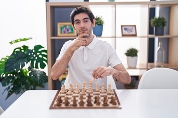 Young hispanic man playing chess sitting on the table serious face thinking about question with hand on chin, thoughtful about confusing idea