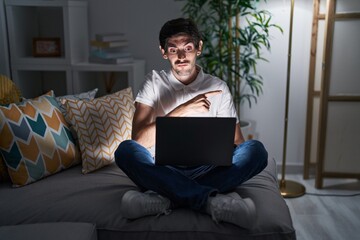 Young hispanic man using laptop at home at night pointing aside worried and nervous with...