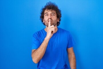 Hispanic young man standing over blue background asking to be quiet with finger on lips. silence...