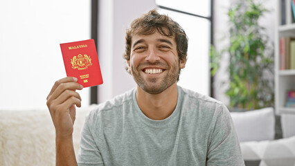 Handsome young man smiling with joy, sitting on a sofa at home, holding his malaysian passport - an...