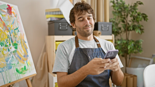 Cheerful young hispanic artist man, blond beard and apron, smiling while typing a message on his smartphone during a break in his vibrant art class at a college studio