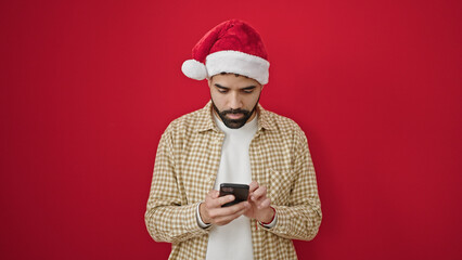 Young hispanic man wearing christmas hat using smartphone over isolated red background