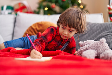 Adorable toddler writing on notebook lying on sofa by christmas tree at home