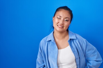 Asian young woman standing over blue background winking looking at the camera with sexy expression,...
