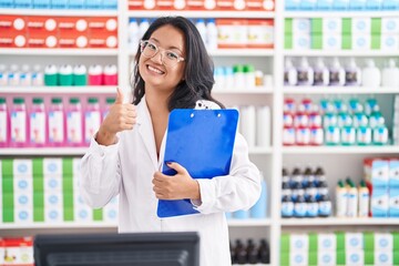Asian young woman working at pharmacy drugstore holding clipboard smiling happy and positive, thumb...