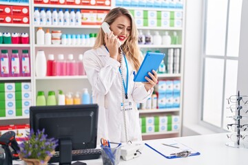 Young beautiful hispanic woman pharmacist using touchpad talking on telephone at pharmacy