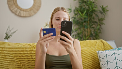 Young blonde woman shopping with smartphone and credit card sitting on sofa at home