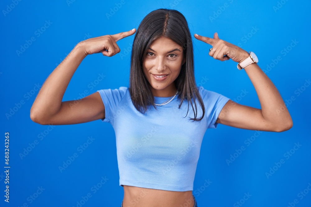 Wall mural brunette young woman standing over blue background smiling pointing to head with both hands finger, 