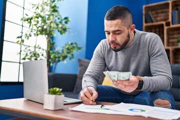 Young latin man writing on document holding dollars at home