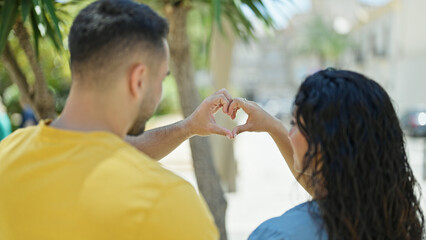 Man and woman couple doing heart gesture standing backwards at park