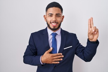 Young hispanic man wearing business suit and tie smiling swearing with hand on chest and fingers up, making a loyalty promise oath