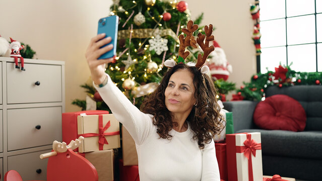 Middle age hispanic woman make selfie by smartphone sitting on floor by christmas tree at home