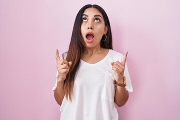 Young arab woman standing over pink background amazed and surprised looking up and pointing with fingers and raised arms.