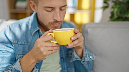Young caucasian man smelling cup of coffee sitting on sofa at home