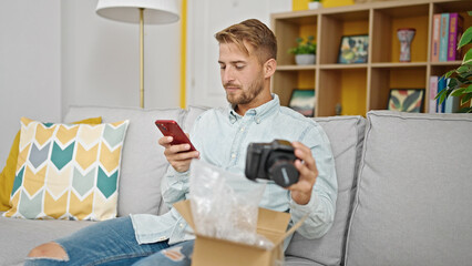 Young caucasian man using smartphone unpacking camera of cardboard box at home
