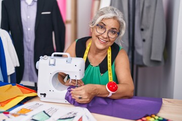 Middle age grey-haired woman tailor smiling confident using sewing machine at tailor shop