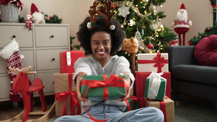 Obraz na płótnie Canvas African american woman wearing reindeer ears holding christmas gift at home