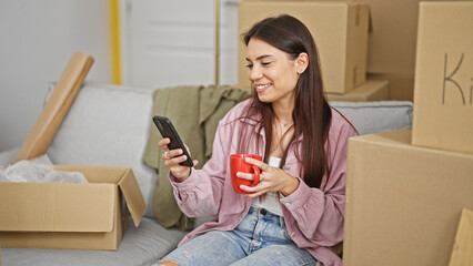 Young beautiful hispanic woman using smartphone drinking coffee at new home