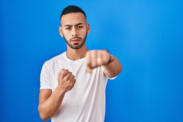 Young hispanic man standing over blue background punching fist to fight, aggressive and angry attack, threat and violence