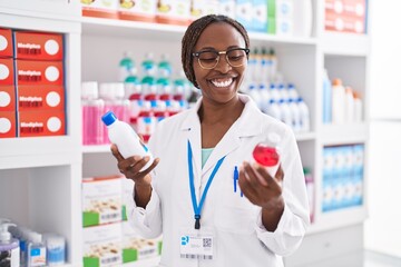 African american woman pharmacist smiling confident holding medication bottles at pharmacy