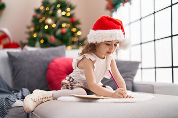 Adorable caucasian girl drawing on notebook sitting on sofa by christmas tree at home