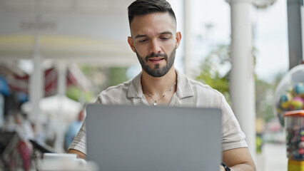 Young hispanic man using laptop sitting on table at coffee shop terrace