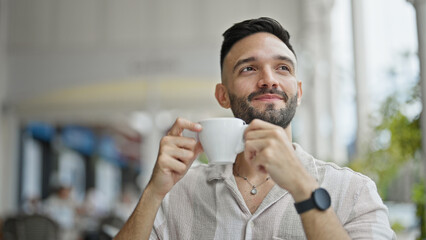 Young hispanic man holding cup of coffee smiling at coffee shop terrace