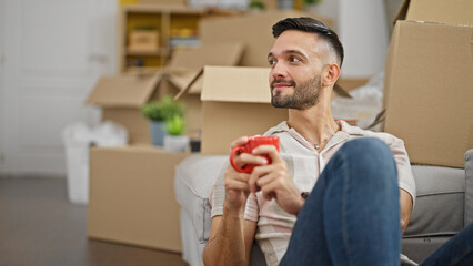 Young hispanic man drinking coffee sitting on floor at new home