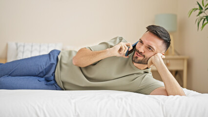 Young hispanic man talking on smartphone lying on bed at bedroom