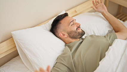 Young hispanic man waking up stretching arms at bedroom