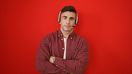 Obraz premium Charming young hispanic man standing firm as call center agent boss, radiating success with relaxed gesture, arms crossed, wearing headset over isolated red wall