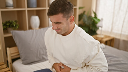 Fototapeta premium Young, attractive hispanic man painfully suffering from stomach-ache in bed, touching his abdomen while sitting awakened in his bedroom's interior