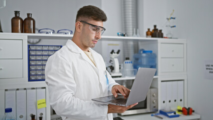 Naklejka na ściany i meble Young hispanic man scientist wearing security glasses using laptop at laboratory