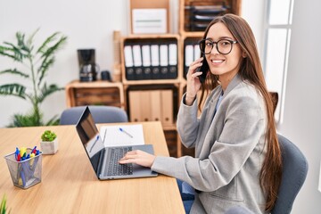 Young beautiful hispanic woman business worker using laptop talking on smartphone at office