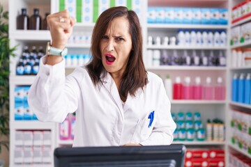 Middle age brunette woman working at pharmacy drugstore angry and mad raising fist frustrated and...