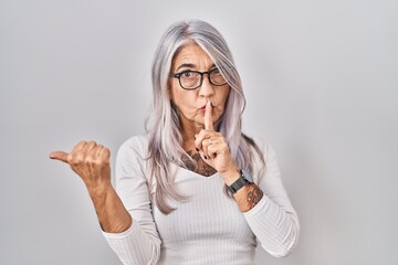 Middle age woman with grey hair standing over white background asking to be quiet with finger on...