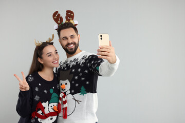 Happy young couple in Christmas sweaters and reindeer headbands taking selfie on grey background....