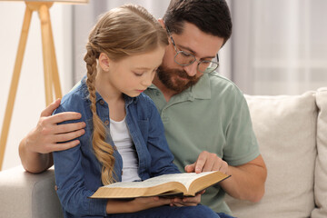 Girl and her godparent reading Bible together on sofa at home