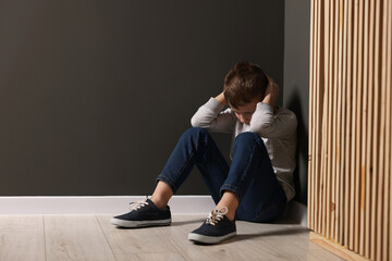 Child abuse. Upset boy sitting on floor near gray wall indoors, space for text