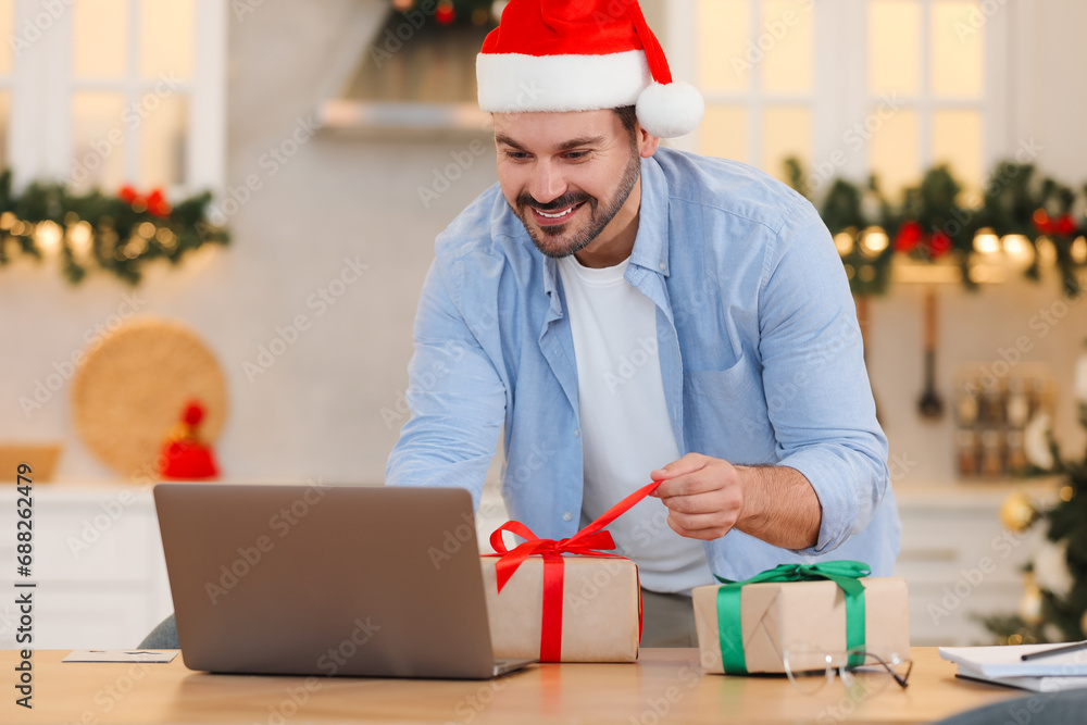 Sticker Celebrating Christmas online with exchanged by mail presents. Smiling man in Santa hat opening gift box during video call on laptop at home