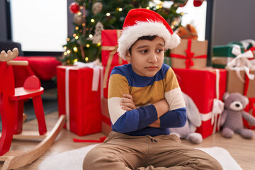 Obraz na płótnie Canvas Adorable hispanic boy sitting on floor by christmas tree with sad expression at home